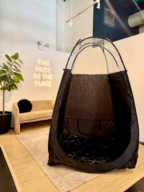 Spray tan tent in a studio beside a glowing This Must Be the Place sign