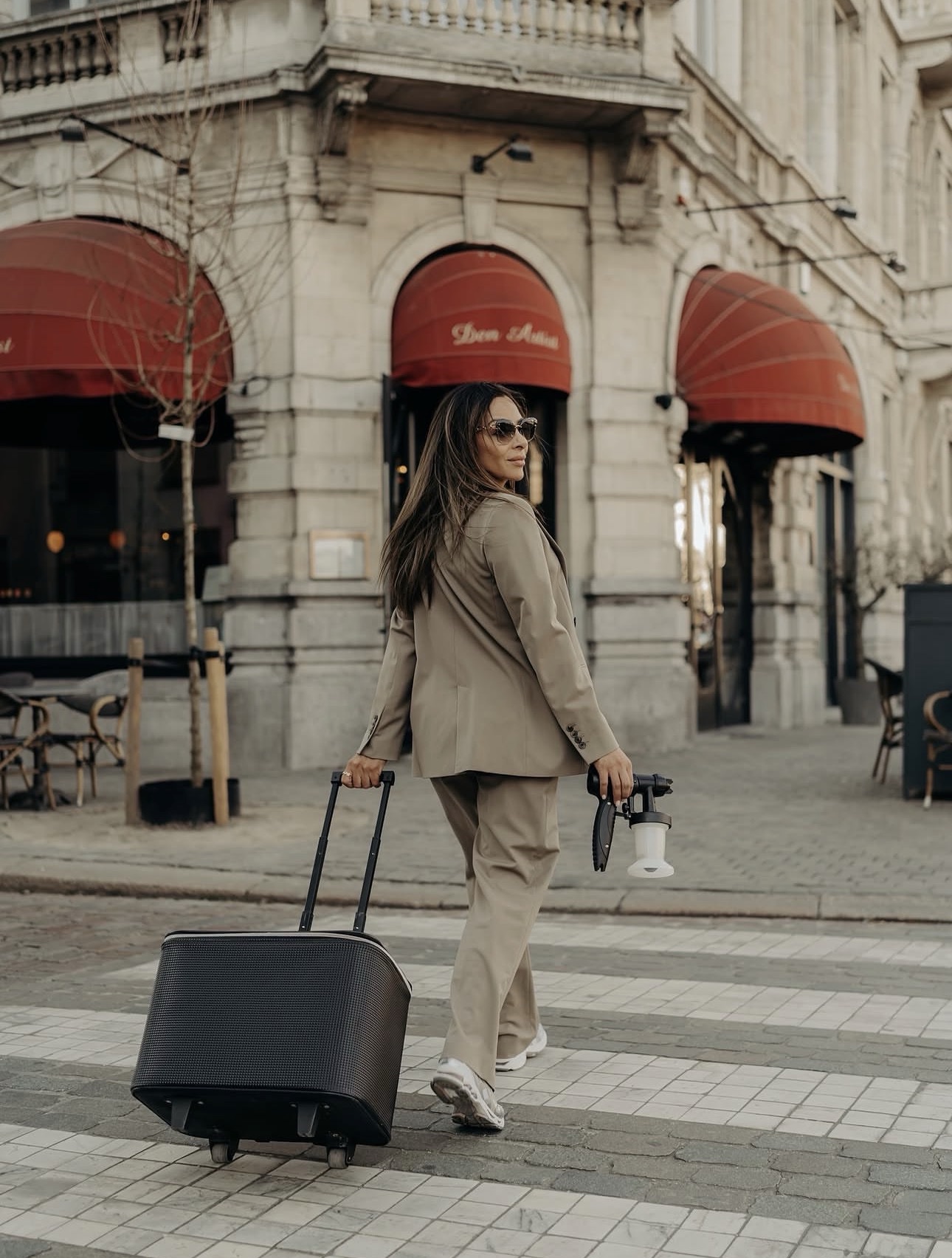 Gigi crossing an Antwerp street in a tailored suit with her mobile spray tan kit