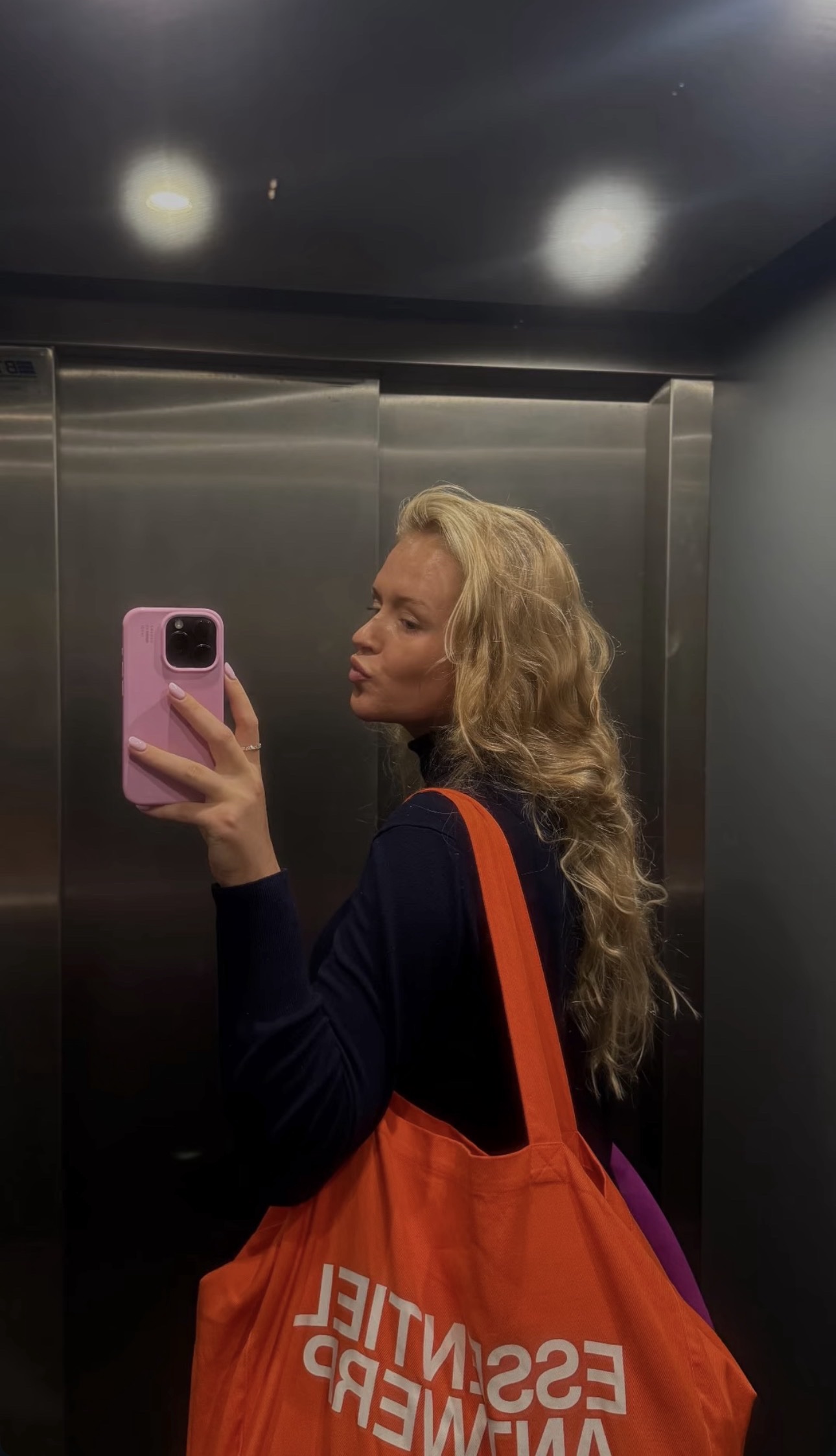 A blonde tour guest taking an elevator mirror selfie with a bright orange tote